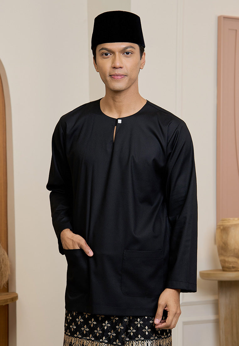 Man wearing a black traditional baju melayu teluk belanga outfit with a matching songkok lipat indoors.
