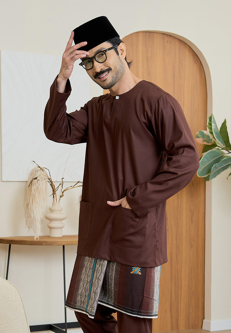 Man wearing a rich brown traditional baju melayu teluk belanga outfit and a songkok lipat indoors with a wooden door and plant in the background