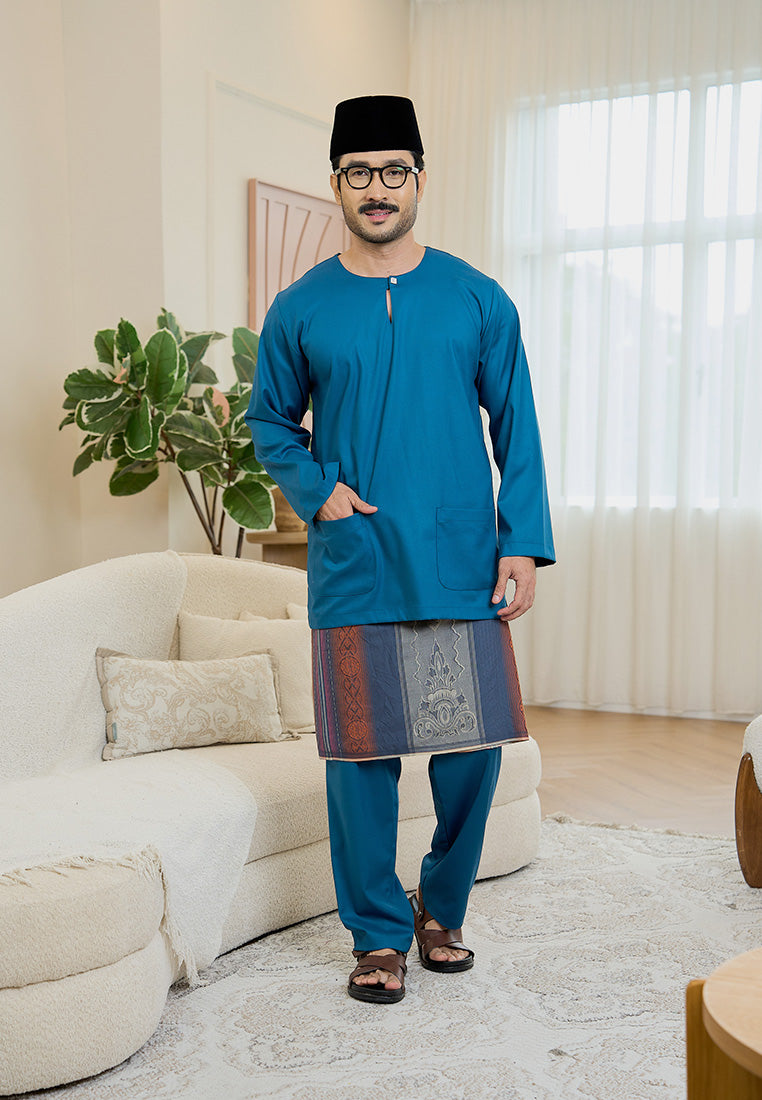 Man wearing a teal blue traditional baju melayu teluk belanga outfit with a black songkok and a samping in a living room.