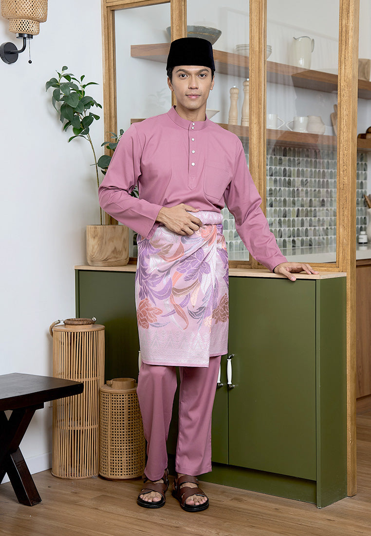 Man wearing a modern baju melayu cekak musang outfit in pink standing in a room with a green cabinet and wooden shelves.
