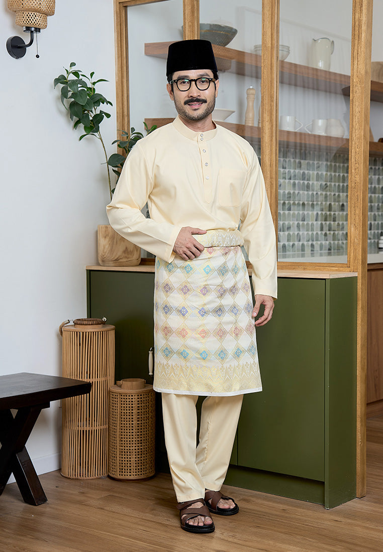 Man wearing a traditional Baju Melayu cekak musang in pastel yellow with samping and songkok standing in a room with wooden furniture and plants.