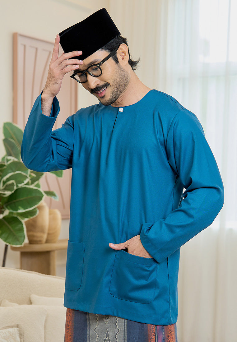Man wearing a teal blue traditional baju melayu teluk belanga outfit with a black songkok lipat indoors.