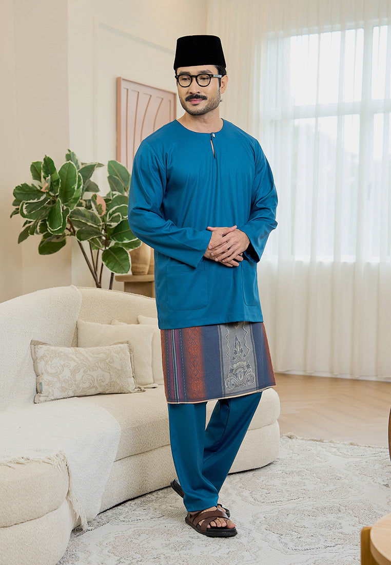 Man with glasses wearing a teal blue traditional baju melayu teluk belanga outfit in a living room setting